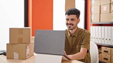 Genç Hispanik iş adamı ofisteki dizüstü bilgisayarla paketleri kontrol ediyor.