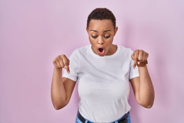 Güzel Afro-Amerikalı kadın pembe arka planda durup parmaklarını göstererek reklamını yapıyor, şaşırmış yüzünü ve ağzını açıyor. 
