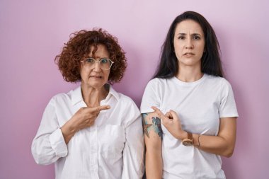 İspanyol anne ve kızı pembe arka plan üzerine gündelik beyaz tişört giyiyorlar. Endişeli ve gergin bir kenara işaret ediyor. İşaret parmağı, endişeli ve şaşırmış ifade. 