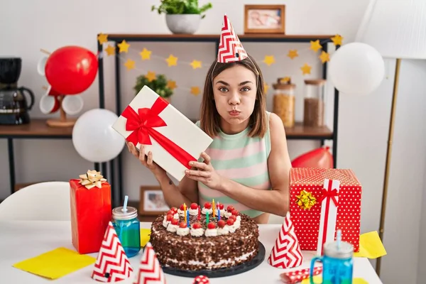 西班牙裔女人拿着蛋糕庆祝生日 脸上带着滑稽的笑脸 手里拿着礼物 嘴被空气吹胀 — 图库照片