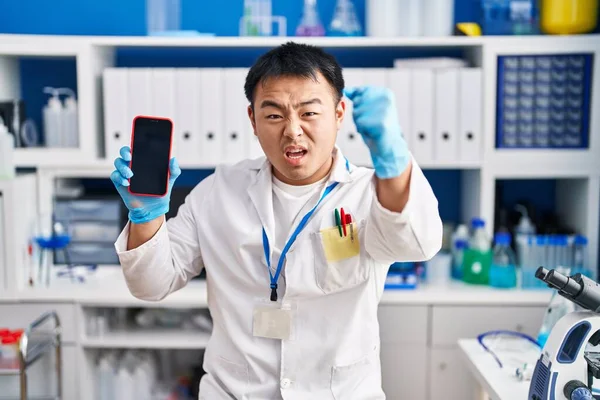 Young chinese man working at scientist laboratory holding smartphone annoyed and frustrated shouting with anger, yelling crazy with anger and hand raised