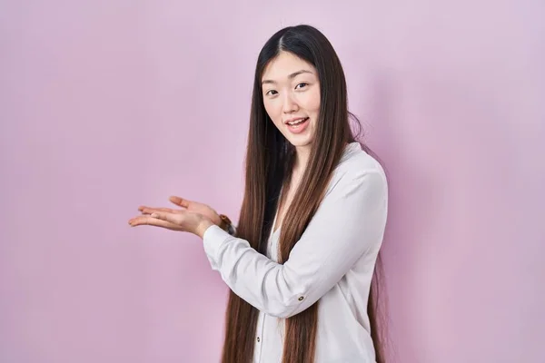 中国の若い女性がピンクの背景の上に立って脇に手を開いてヤシの木は コピースペースを示す 広告笑顔興奮幸せな提示 — ストック写真