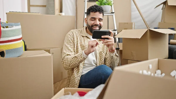 Young hispanic man watching video on smartphone sitting on floor at new home