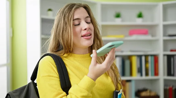 在图书馆大学 年轻的金发女学生拿着书本 用智能手机传送语音讯息 — 图库照片