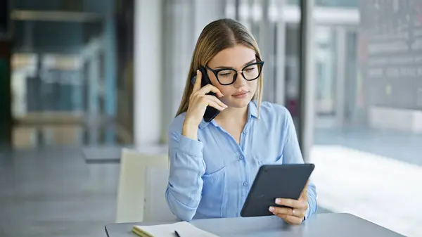 Young blonde woman business worker using touchpad talking on smartphone at office