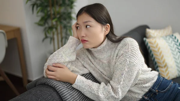 若いです中国人女性座っていますソファに深刻な表現とともに自宅 — ストック写真