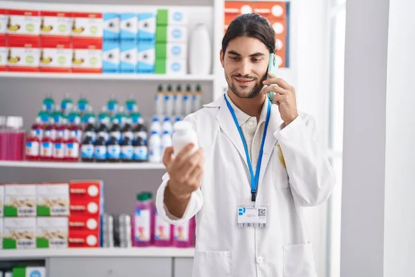 Joven Farmacéutico Hispano Sosteniendo Botella Pastillas Hablando Teléfono Inteligente Farmacia —  Fotos de Stock