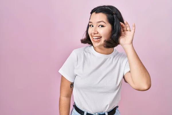 ピンクの背景にカジュアルな白いTシャツを着た若いヒスパニック系の女性が 噂やゴシップに耳を傾け 耳を傾けながら手で笑っています 聴覚障害の概念 — ストック写真