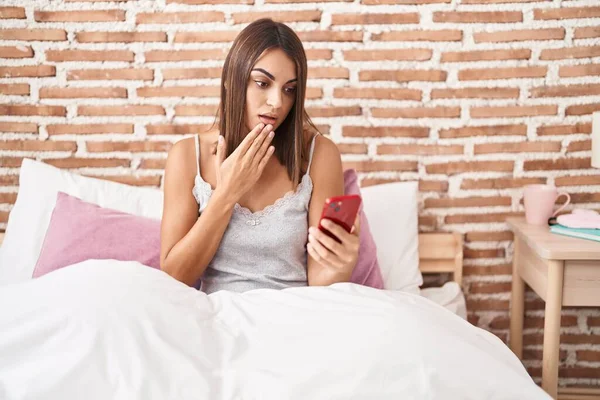 年轻的惊慌失措的女人用智能手机坐在床上 用手捂住嘴 惊恐万分 生怕犯错 惊讶的表情 — 图库照片