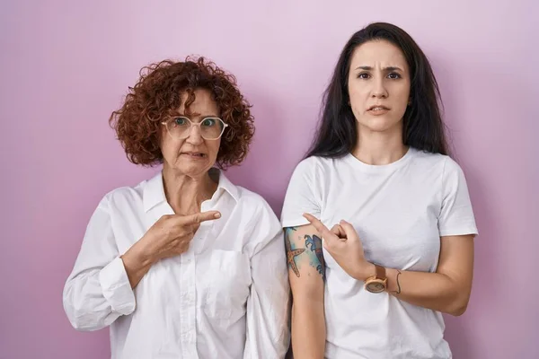 ヒスパニック系の母親と娘身に着けていますカジュアルホワイトTシャツ上のピンクの背景指摘脇心配と神経で前指 懸念と驚きの表現 — ストック写真