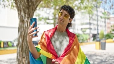 Gökkuşağı bayrağı takan İspanyol genç adam parkta akıllı telefondan selfie çekiyor.