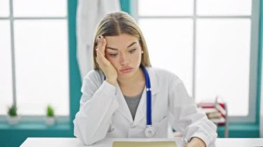 Genç sarışın kadın doktor, klinikte masada yorgun oturuyor.