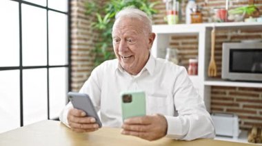 Yemek odasındaki masada oturan yaşlı gri saçlı adam akıllı telefon kullanıyor.