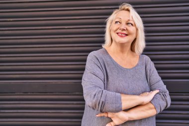 Orta yaşlı sarışın kadın gülümsüyor. Kollarını kavuşturup caddede duruyor.