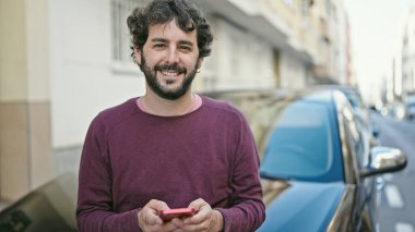 Young hispanic man using smartphone leaning on car at street