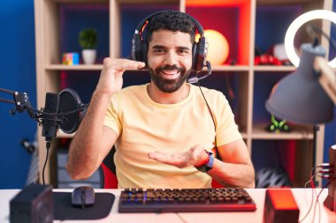 Sakallı, video oyunu oynayan, kulaklıklı, büyük ve büyük boy işareti gösteren İspanyol bir adam. Ölçü sembolü. Kameraya bakıp gülümsüyor. ölçüm kavramı. 