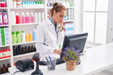 Young beautiful hispanic woman pharmacist talking on telephone using computer at pharmacy