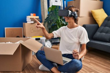 Young beautiful hispanic woman playing video game using virtual reality glasses sitting on floor at new home