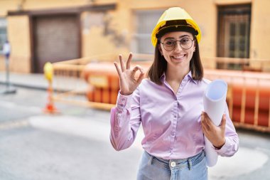 Hispanic girl wearing architect hardhat at construction site doing ok sign with fingers, smiling friendly gesturing excellent symbol 