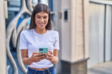 Genç, güzel, İspanyol bir kadın sokakta akıllı telefon kullanarak gülümsüyor.