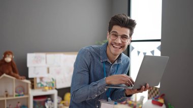 Young hispanic man preschool teacher using laptop standing at kindergarten