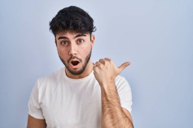 Hispanic man with beard standing over white background surprised pointing with hand finger to the side, open mouth amazed expression. 
