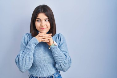 Mavi arka planda dikilen genç İspanyol kadın. Gergin ve heyecanlı. Elleri çenesinde yan tarafa bakıyor. 