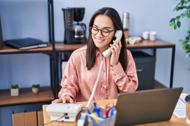 Genç İspanyol iş kadını laptopunu kullanıyor ve ofisteki akıllı telefondan konuşuyor.