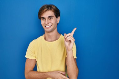 Mavi arka planda duran genç adam yüzünde kocaman bir gülümsemeyle, eli ve parmağı kameraya bakıyor.. 