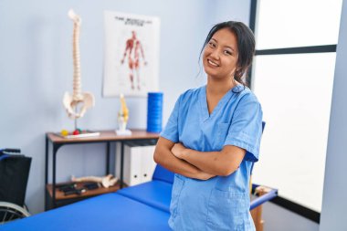 Young chinese woman wearing physiotherapist uniform standing with arms crossed gesture at rehab clinic