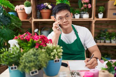 Young chinese man florist talking on smartphone writing on notebook at flower shop