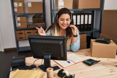 Young hispanic woman working at small business ecommerce wearing headset smiling happy pointing with hand and finger to the side 