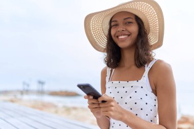 Young african american woman wearing summer hat using smartphone at seaside