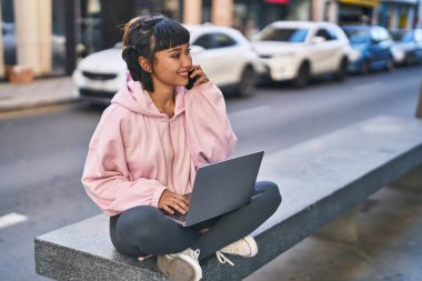 Dizüstü bilgisayar kullanan genç bir kadın akıllı telefonla konuşuyor. Sokakta bankta oturuyor.
