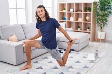 Young hispanic man stretching leg sitting on floor at home