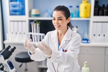 Young beautiful hispanic woman scientist holding test tubes with flowers at laboratory