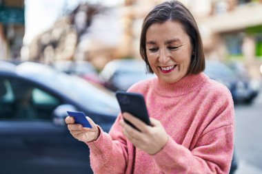 Sokakta akıllı telefon ve kredi kartı kullanan orta yaşlı bir kadın.