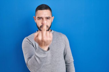 Hispanic man standing over blue background showing middle finger, impolite and rude fuck off expression 