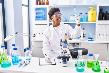 Bilim adamı üniforması giyen Afrikalı Amerikalı bir kadın laboratuarda elinde test tüpleriyle panoya yazı yazıyor.