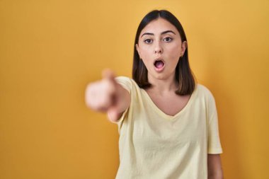 İspanyol kız sarı arkaplan üzerine gündelik tişört giyiyor. Parmakları önde, ağzı açık, şaşırmış, ön tarafta bir şey var. 