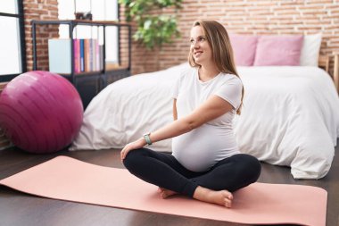 Yatak odasındaki yoga minderinde oturan hamile bir kadın hazırlık egzersizi yapıyor.