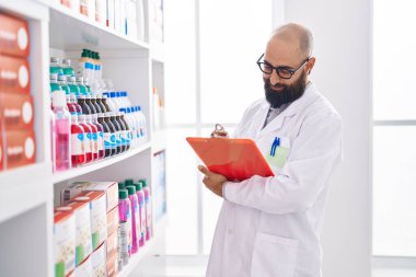 Young bald man pharmacist smiling confident writing on clipboard at pharmacy