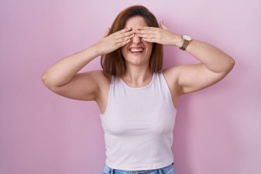 Esmer kadın pembe arka planda duruyor. Gözleri kaplıyor. Gülümseyen neşeli ve komik ellerle. kör kavram. 