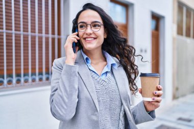 Genç İspanyol kadın yönetici kahve içiyor ve caddede akıllı telefondan konuşuyor.