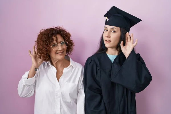 卒業キャップと儀式用のローブを身に着けているヒスパニック系の母親と娘は噂やゴシップへの聴聞会を耳に手で笑みを浮かべて 聴覚障害の概念 — ストック写真