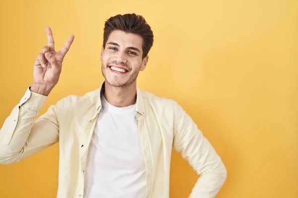 Sarı Arka Planda Dikilmiş Gülümseyen Genç Spanyol Adam Parmaklarını Zafer — Stok fotoğraf