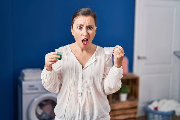衝撃的な顔で洗濯洗剤と洗濯粉末を保持している中年のパニック女性 懐疑的で皮肉を探して オープン口で驚いて — ストック写真
