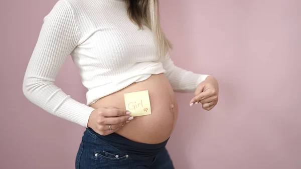 若いです妊娠女性指しています女の子リマインダー紙上の腹上の隔離されたピンクの背景 — ストック写真