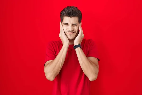 大きな音楽の音にいらいらする表情で指で耳を覆う赤い背景の上に立つ若いヒスパニック系の男 聴覚障害の概念 — ストック写真