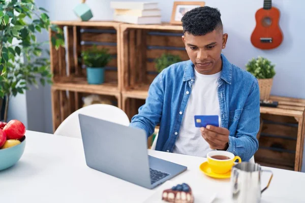 Young latin man using credit card using laptop having breakfast at home